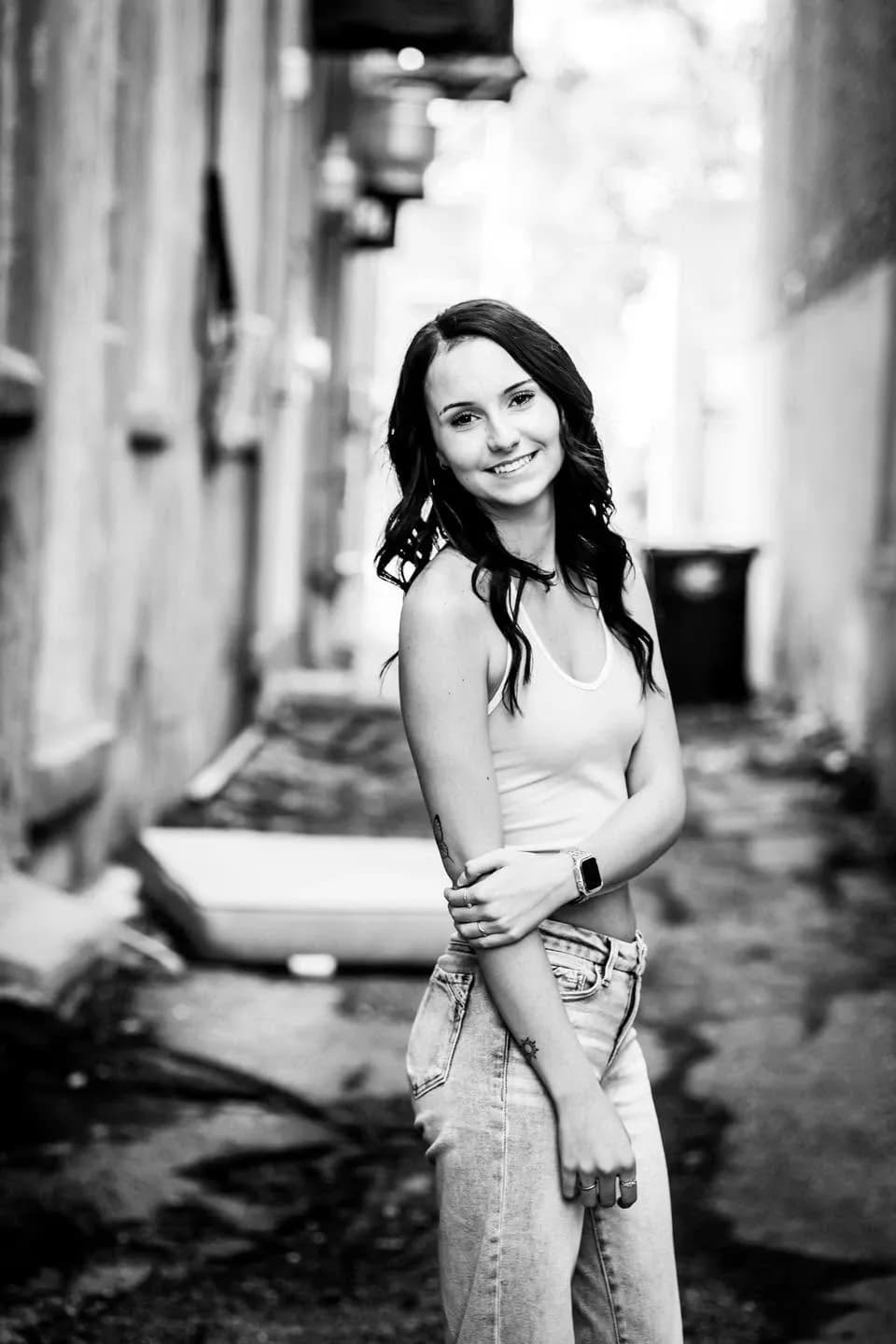 Black-and-white portrait of a smiling young woman in a tank top and jeans in a narrow alley