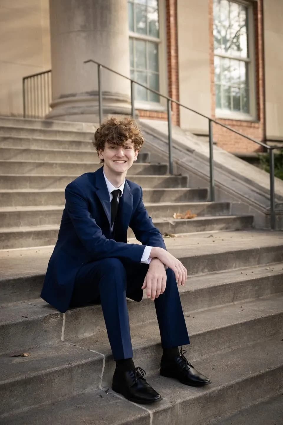 Young man in a navy suit and tie smiling while seated on stone steps outside a columned brick building