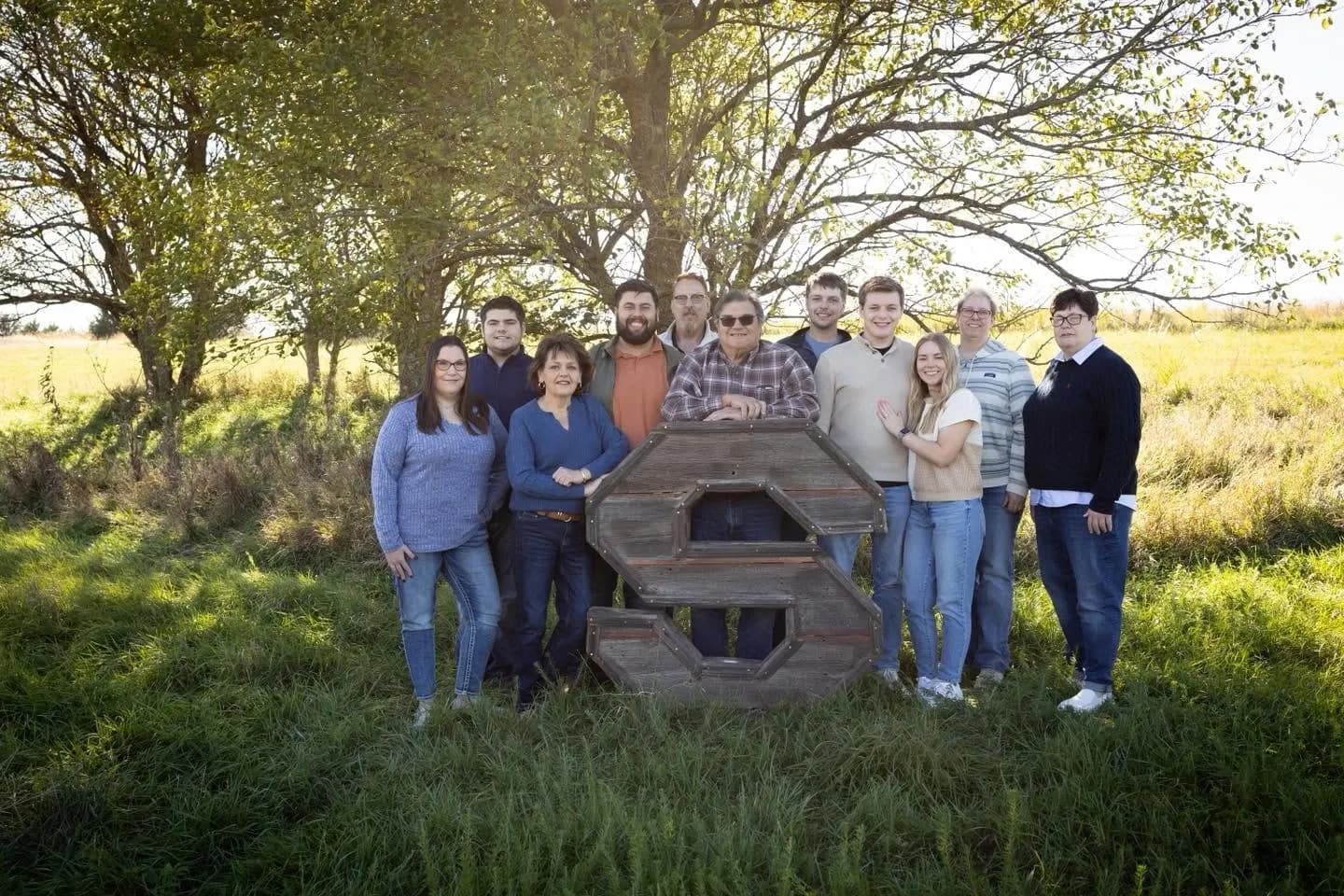 Multigenerational family posing outdoors around a large wooden letter S in a grassy field at golden hour