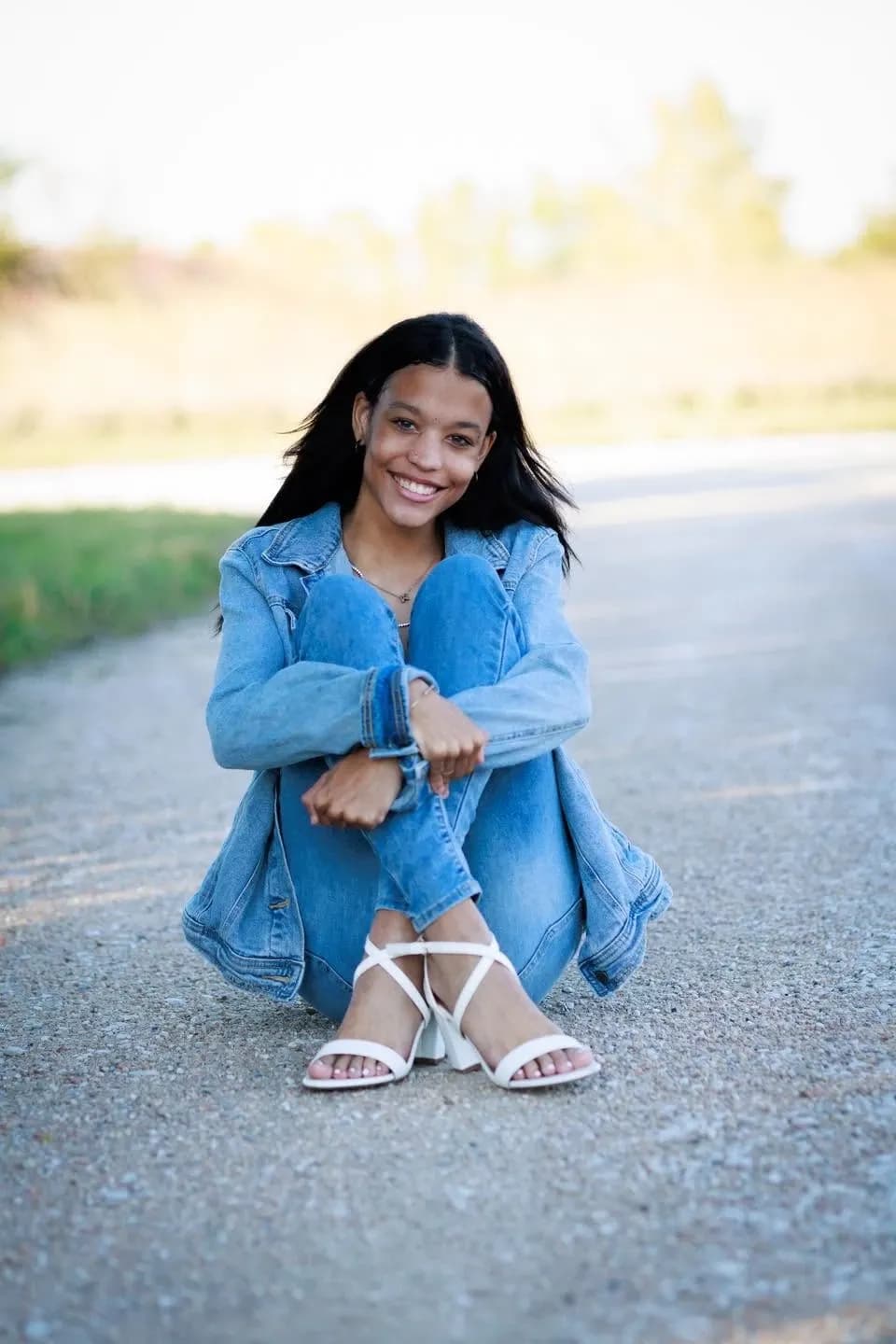Smiling young woman in a denim jacket and jeans sitting on a country road with her knees drawn up