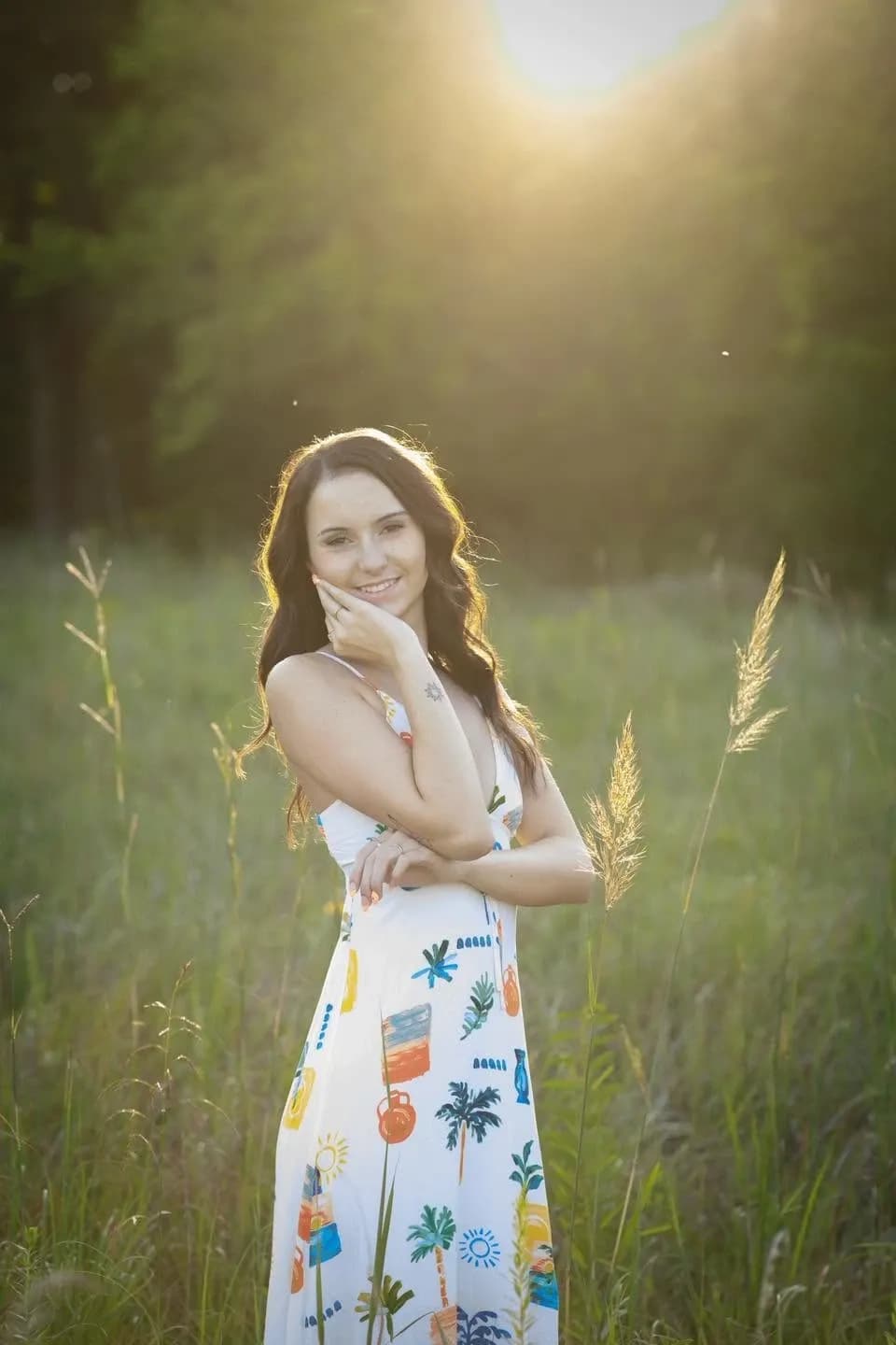 Young woman in a white floral-print dress smiling in a sunlit grassy field at golden hour