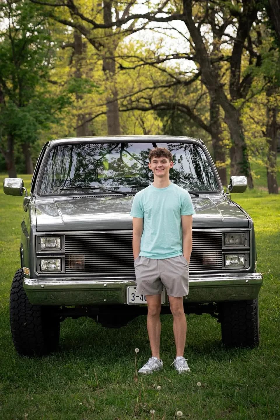Smiling teen in a teal shirt standing with hands in pockets before a lifted vintage pickup in a wooded field