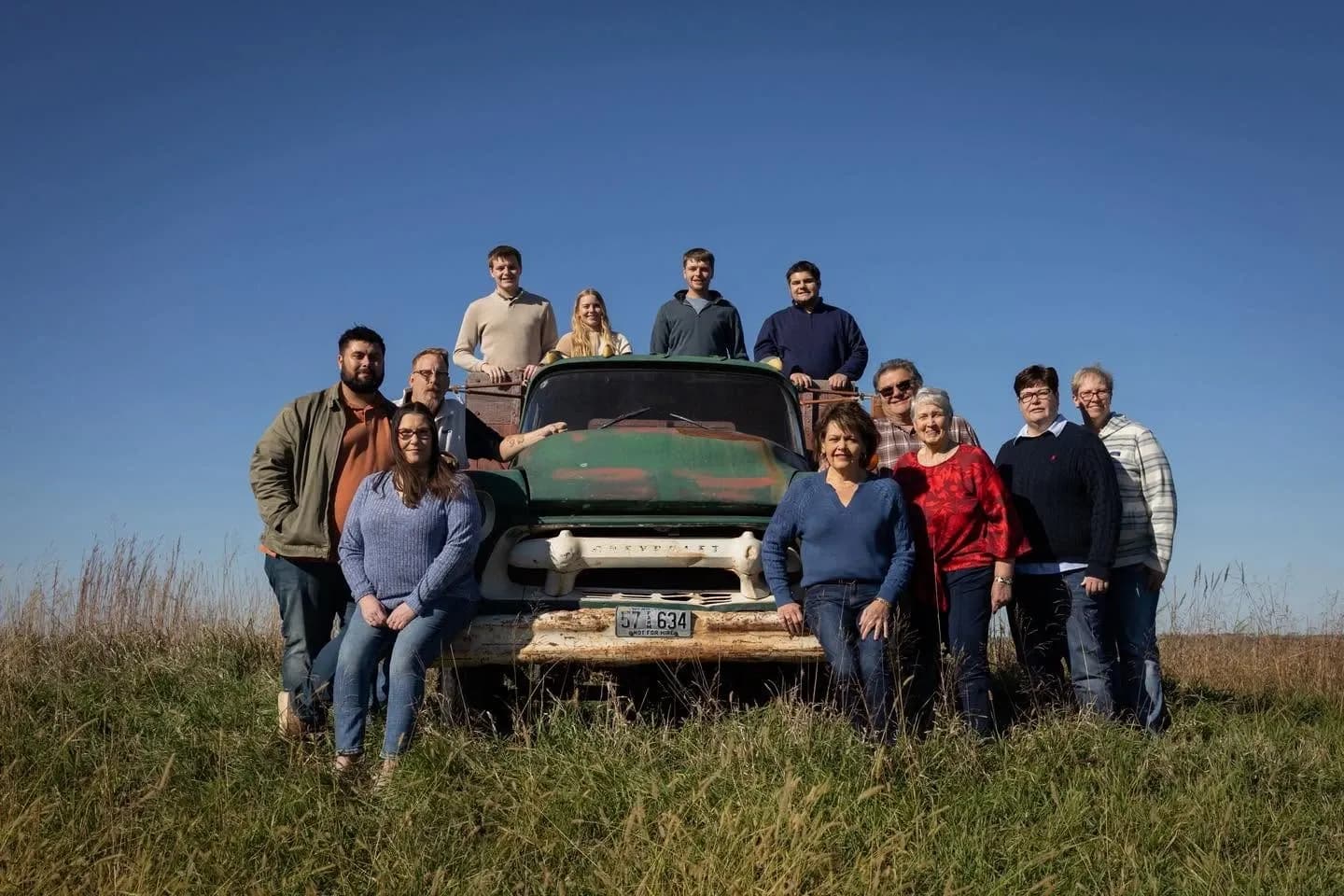 Large multigenerational family gathered around a weathered vintage green truck in a sunlit prairie field