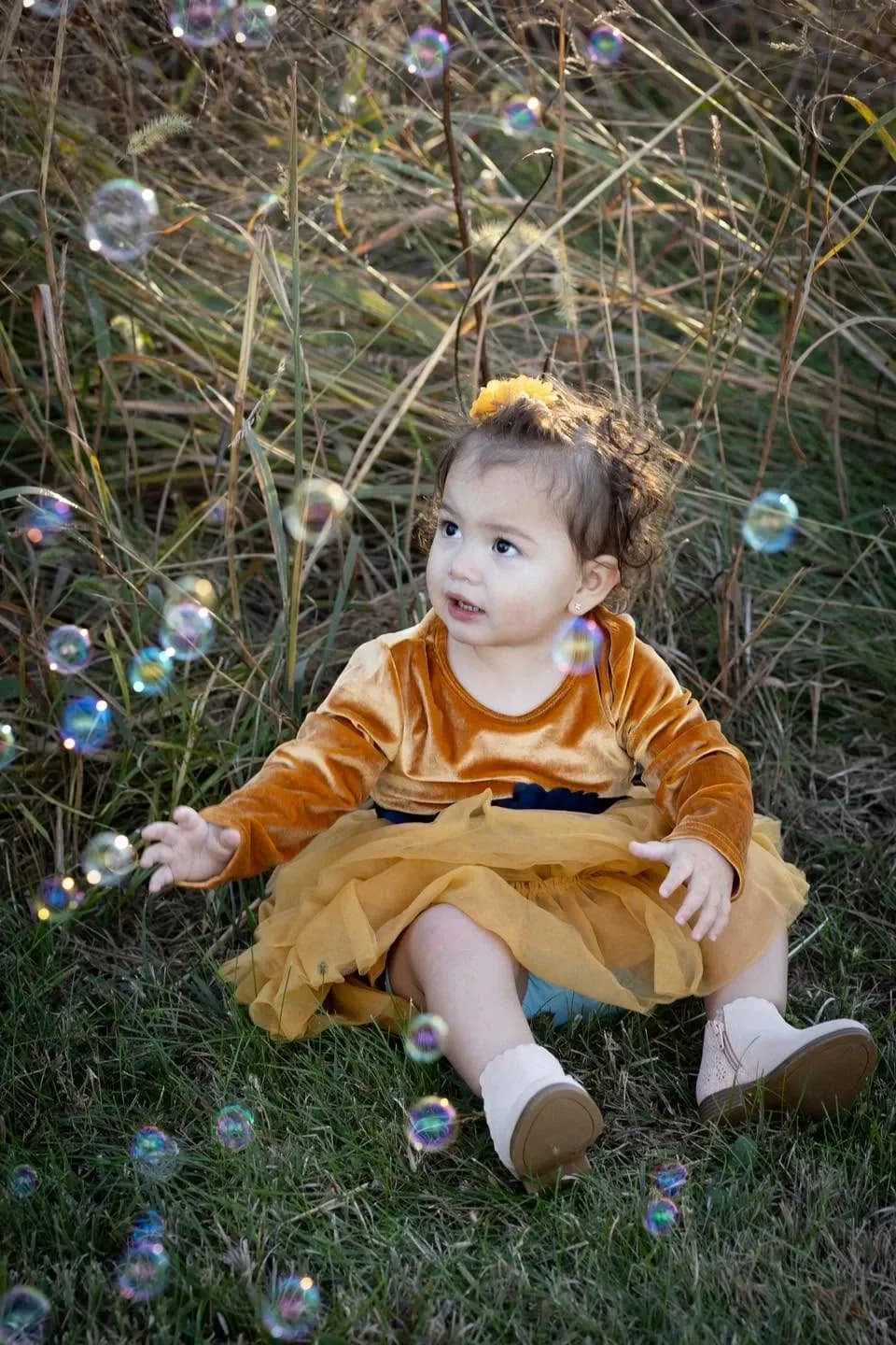 Toddler in a mustard velvet dress reaching toward floating soap bubbles in a grassy field at golden hour