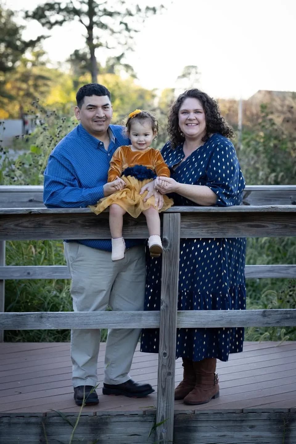 Family of three in coordinated navy and gold outfits on a wooden bridge, a toddler seated on the railing