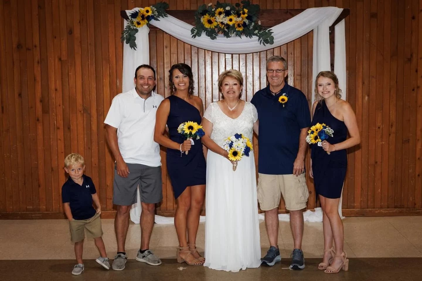 A bride holding a sunflower bouquet stands with five family members under a sunflower-decorated arch