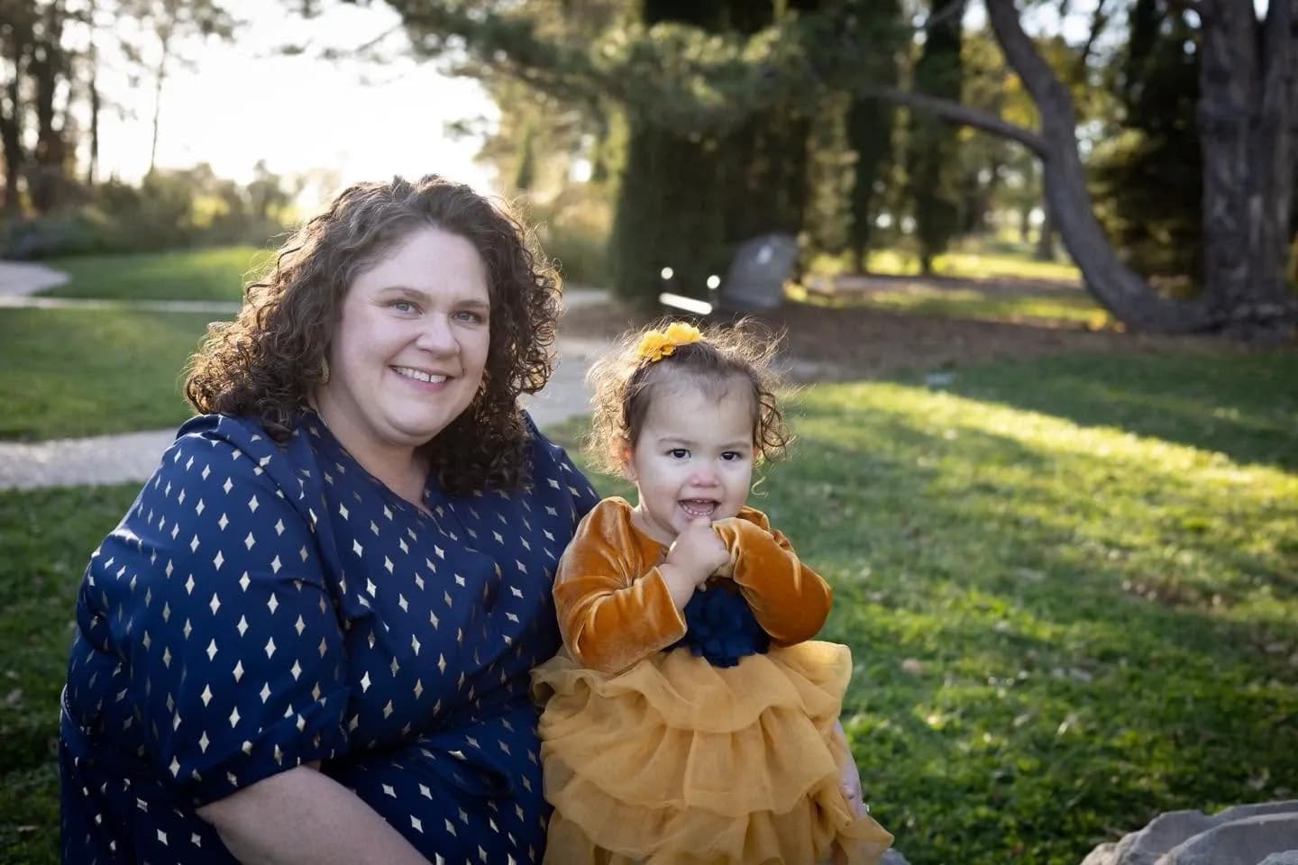 A woman in navy sits beside a toddler in a gold ruffled dress in a sunlit park at golden hour
