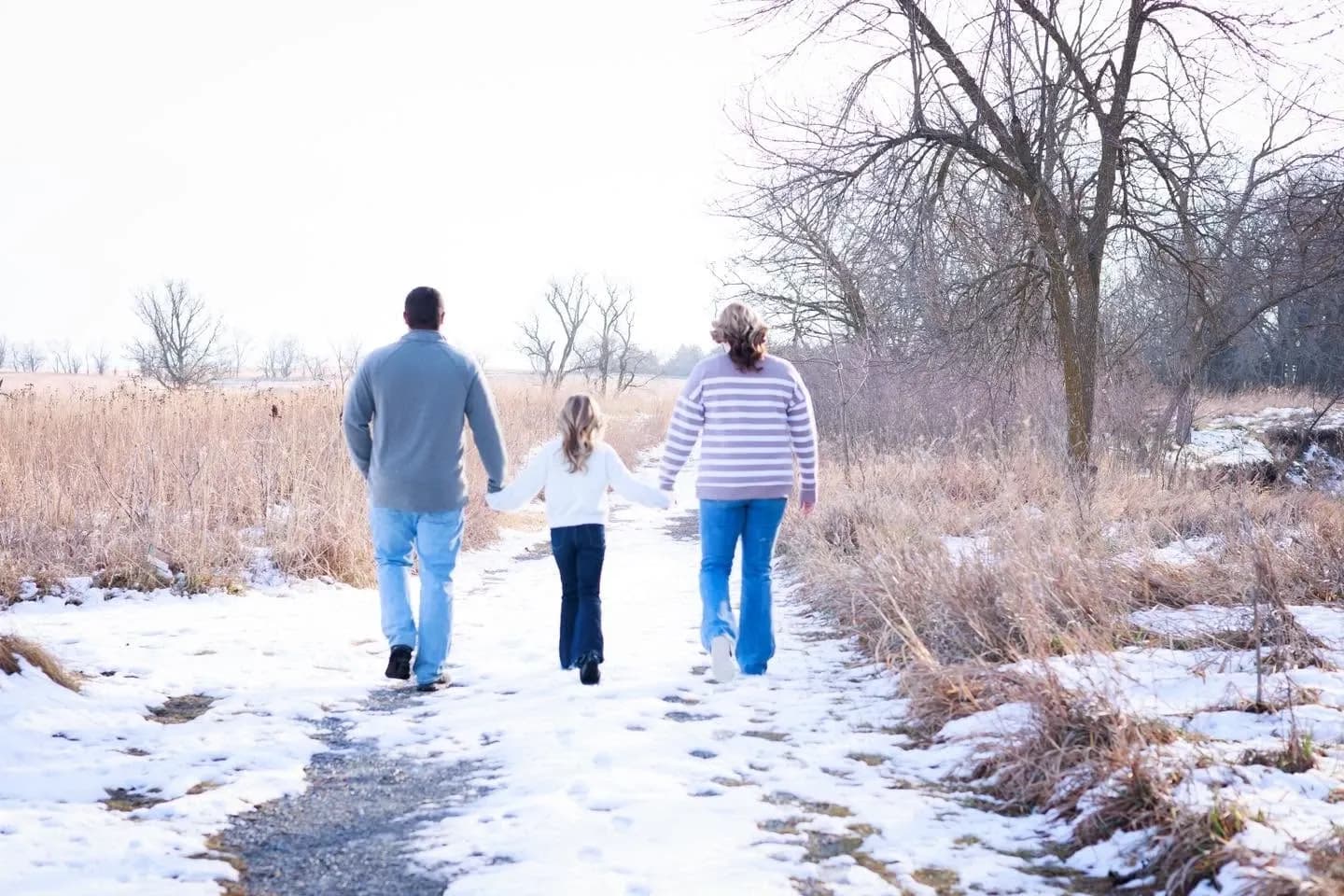 Family of three walking hand in hand down a snowy path among bare trees and tall winter grass