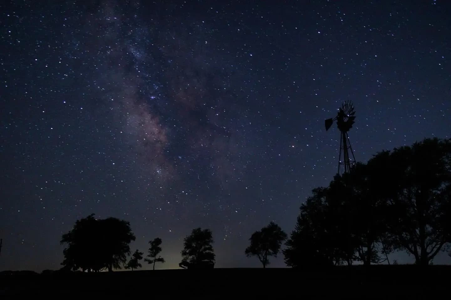 The Milky Way over a rural night sky with a silhouetted farm windmill and trees