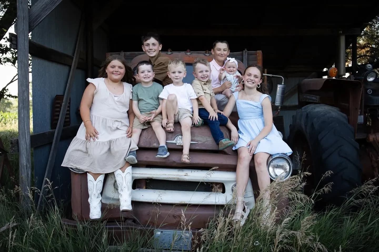 Eight children smiling together on the hood of a vintage rusty truck in a rural barn setting