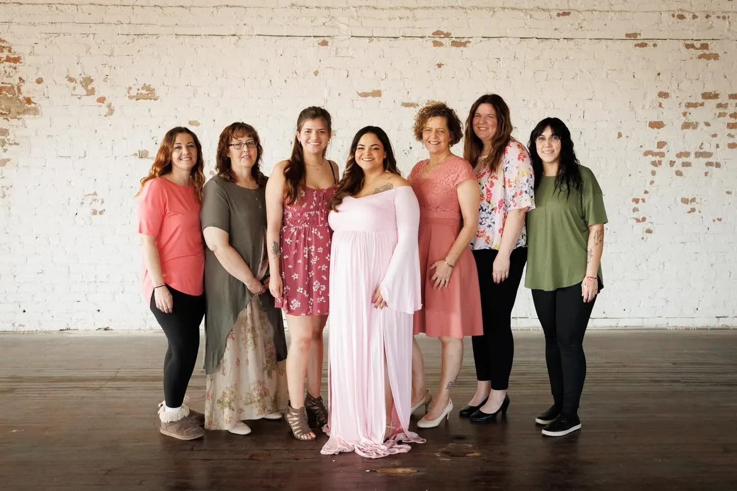 Seven women posing together against a whitewashed brick wall, the central woman in a long flowing pink gown