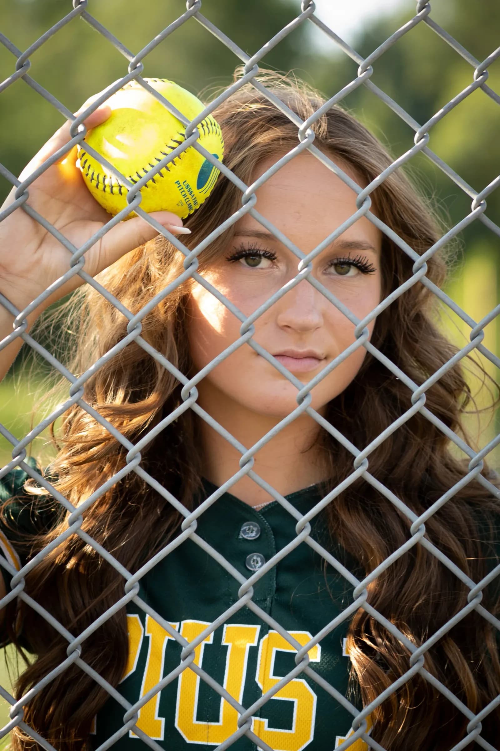 Girl holding softball looking thru chain linkmfence